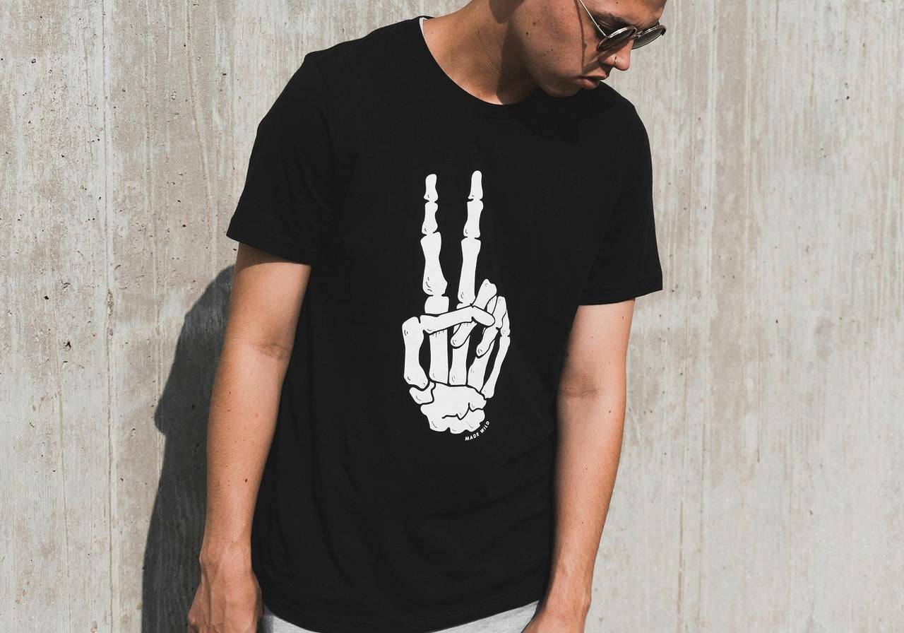 A young man in sunglasses and a black t-shirt with a skeleton hand peace sign design against a concrete wall.
